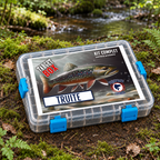 Target Box Truite mouchetée de Target Baits Leurres, coffret fermé à attaches bleues sur une berge de terre et de mousse près d’un ruisseau.